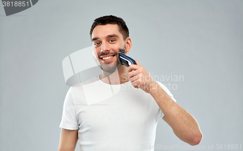 Image of smiling man shaving beard with trimmer over gray