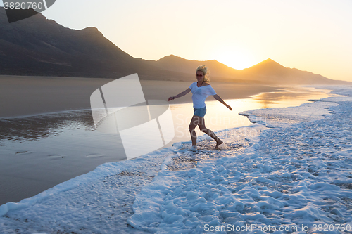 Image of Lady enjoying running from waves on sandy beach in sunset.