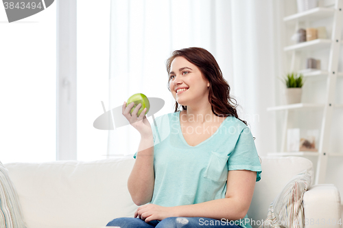 Image of happy plus size woman eating green apple at home