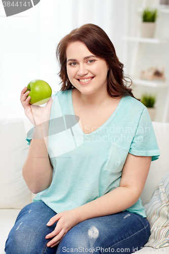 Image of happy plus size woman eating green apple at home