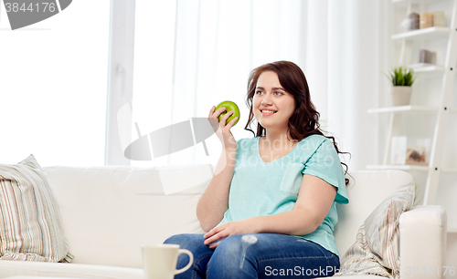 Image of happy plus size woman eating green apple at home