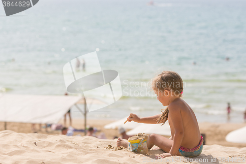 Image of The little five year old girl sitting on the beach and playing with sand and toys on the background of the sea