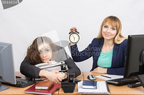 Image of The girl in the office with a smile shows the time on the clock next to the exhausted colleague