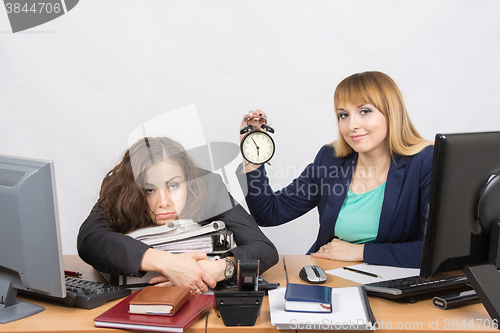Image of Two girls in the office at the end of the day, one with a smile, holding a clock, another weary lies on folders