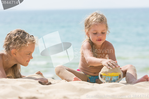 Image of Two girls on the beach on a sunny day, playing with sand and a bucket on a background of the sea