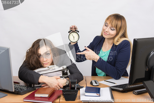 Image of The girl in the office with a smile, holding a clock and looking at the tormented colleague