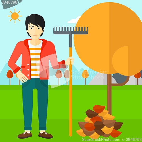 Image of Man with rake standing near tree and heap of autumn leaves.