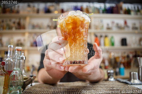 Image of Bartender is standing in pub? holding cocktail and giving glass forward. Focus on beverage.