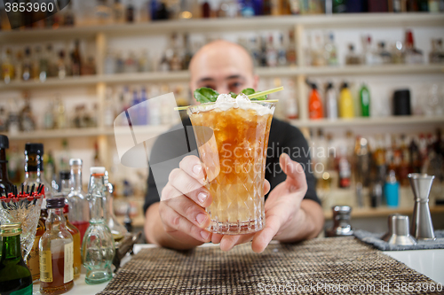 Image of Bartender is standing in pub? holding cocktail and giving glass forward. Focus on beverage.
