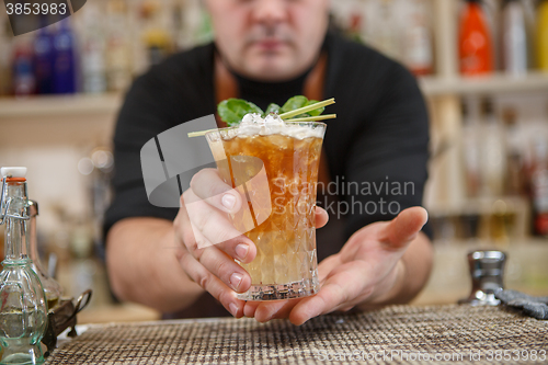 Image of Bartender is standing in pub? holding cocktail and giving glass forward. Focus on beverage.