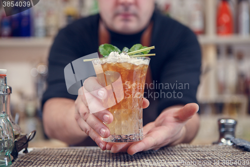 Image of Bartender is standing in pub? holding cocktail and giving glass forward. Focus on beverage.