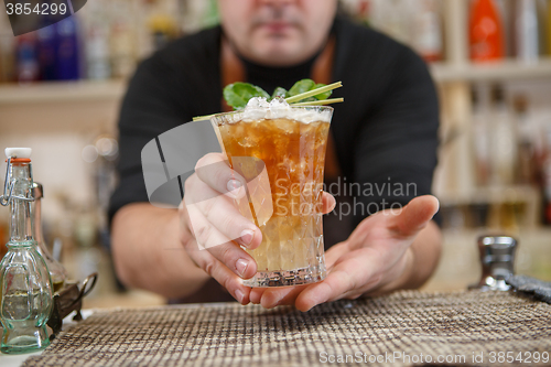 Image of Bartender is standing in pub? holding cocktail and giving glass forward. Focus on beverage.