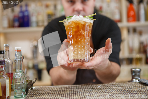 Image of Bartender is standing in pub? holding cocktail and giving glass forward. Focus on beverage.