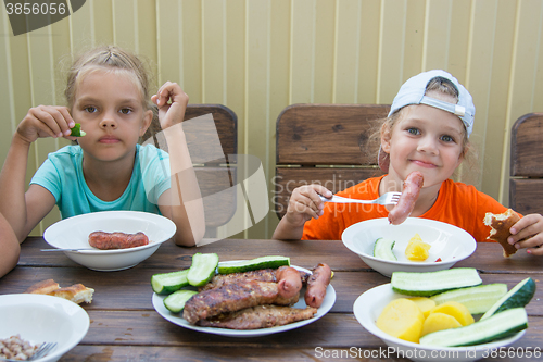 Image of Two little girls at a wooden table in nature eating grilled sausages