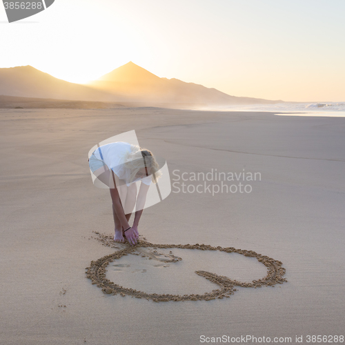 Image of Lady drawing heart shape in sand on beach.