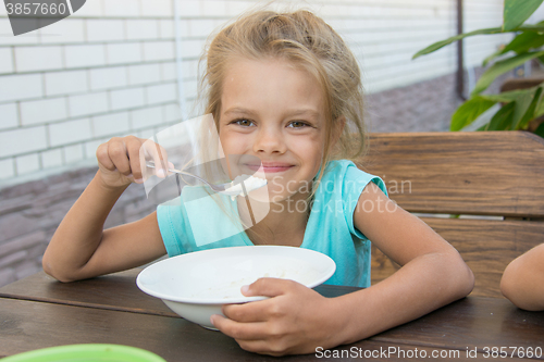 Image of Satisfied six year old girl at the table in the courtyard eating porridge