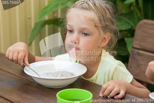 Image of A girl of four years at a table outdoors thoughtfully eating porridge