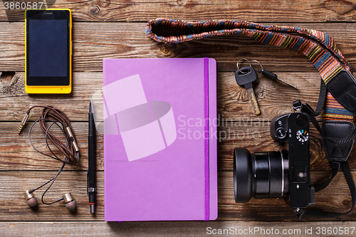 Image of Overhead view of travel gear placed on wooden table. Mobile phone, earplugs, violet sketchbook, pencil, camera and purse. Flat lay top view.