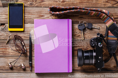 Image of Overhead view of travel gear placed on wooden table. Mobile phone, earplugs, violet sketchbook, pencil, camera and purse. Flat lay top view.