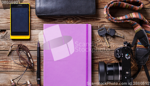 Image of Overhead view of travel gear placed on wooden table. Mobile phone, earplugs, violet sketchbook, pencil, camera and purse. Flat lay top view.