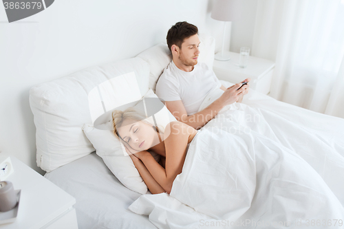 Image of man texting message while woman is sleeping in bed