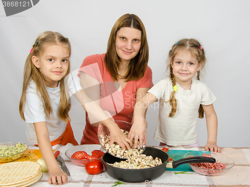 Image of Two little daughter at the kitchen table helping her mother pour chopped mushrooms from the plate to the pan