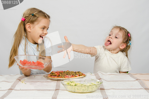 Image of Two little girls at a table prepared pizza. One with a whimsical view stretches a hand to a plate with tomatoes, which takes the other with a smile