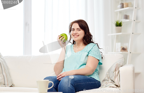 Image of happy plus size woman eating green apple at home