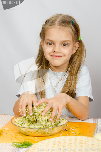 Image of The girl at the kitchen table with a slight smile mixes hands in a bowl the grated cheese with herbs
