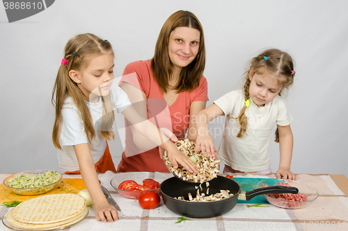 Image of Two little girls at the kitchen table with a zeal to help her mother pour the mushrooms from the plate to the pan