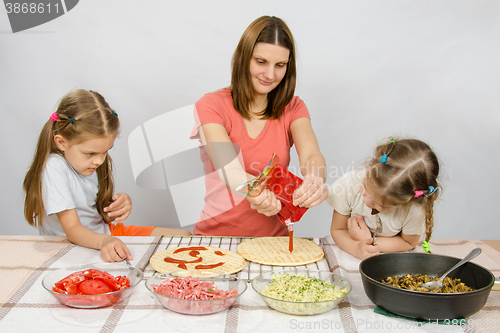 Image of Two little girls enthusiastically watched as mum pours ketchup basis for pizza