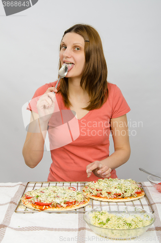 Image of Girl sitting at a table in front of the two has not yet baked pizzas and licks the spoon