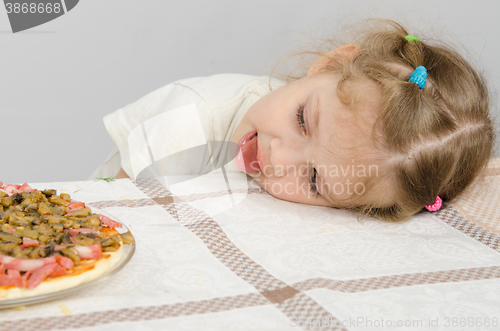 Image of Little girl with protruding tongue rested her head on the table and looks at the pizza