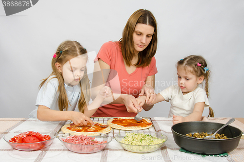 Image of Mom helps younger daughter spread ketchup on a pizza, the eldest daughter, she is preparing a second pizza