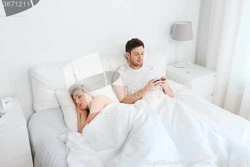 Image of man texting message while woman is sleeping in bed