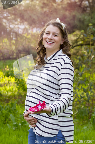 Image of pregnant woman holding baby shoes in her hands in a park