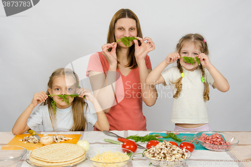 Image of Young housewife with two daughters having fun holding sprig of parsley as a mustache at the kitchen table when sharing cooking