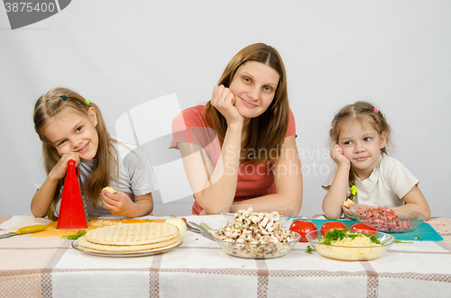 Image of Satisfied mother with two daughters sitting resting his head in his hands at the table with the products for pizza