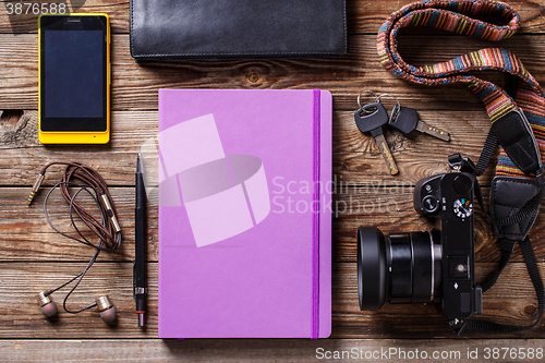 Image of Overhead view of travel gear placed on wooden table. Mobile phone, earplugs, violet sketchbook, pencil, camera and purse. Flat lay top view.