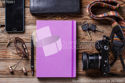 Image of Overhead view of travel gear placed on wooden table. Mobile phone, earplugs, violet sketchbook, pencil, camera and purse. Flat lay top view.