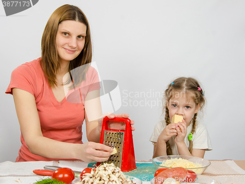 Image of The girl at the kitchen table t cheese grater sitting next to a five-year daughter and eats cheese