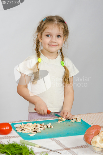 Image of Little girl stands at the kitchen table, with a smile, looking into the frame and slice the mushrooms with a knife