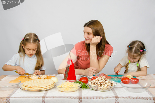 Image of Satisfied mother watching her little daughter to help her cut products at the kitchen table