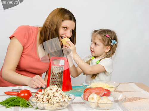 Image of Six-year girl with pigtails giving mom a bite out of a piece of cheese at the kitchen table where they cook together