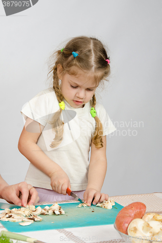 Image of Little girl stands at the kitchen table and helps chop the mushrooms with a knife