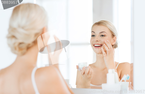 Image of happy woman applying cream to face at bathroom