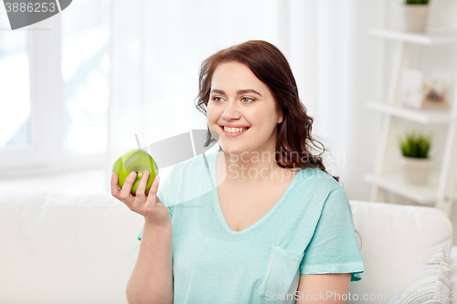 Image of happy plus size woman eating green apple at home