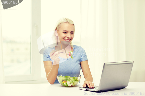 Image of smiling woman with laptop eating salad at home