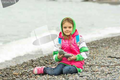 Image of Seven-year girl sitting on a pebble beach in the warm clothes and looks in the frame