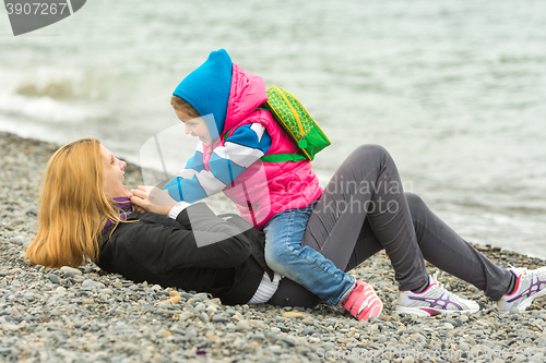 Image of Five-year girl in warm clothes sits astride the mother who is lying on the pebble beach and sea fun looking at each other